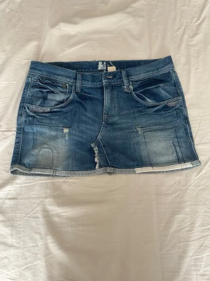 Blå jeanskjol med slitningar - Säljer en blå jeanskjol med rå kant och slitna detaljer. Kjolen har klassiska fem fickor, dragkedja och knapp framtill samt en liten läderpatch bak i midjan. Perfekt för en avslappnad och trendig look.