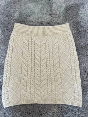 Beige stickad kjol  - Säljer en beige stickad kortkjol från Amisu/New Yorker med snyggt kabelstickat mönster och bred resår i midjan. Kjolen är mjuk och har en klassisk, ribbad kant upptill och nertill. Perfekt för dig som gillar mysiga och trendiga plagg!