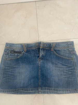Blå jeanskjol från Denim It - Säljer en klassisk blå jeanskjol från Denim It med fem fickor, bälteshällor och knappstängning fram. Kjolen har en kort modell och slitna detaljer för en cool, avslappnad look. Perfekt till sneakers eller boots.