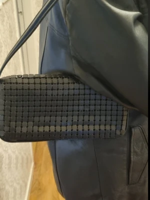 Svart axelväska med rutmönster - Snygg svart axelväska med rektangulär form och rutmönstrad yta gjorde av metal . Väskan har ett kort axelband och minimalistisk design som ger en modern vibe. Perfekt för dig som gillar stilrena accessoarer med en twist.