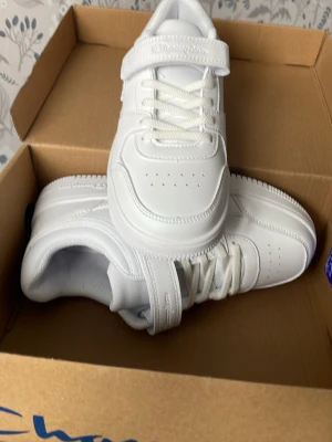 Vita sneakers från Champion - Snygga helvita sneakers från Champion med klassisk låg profil. Skorna har snörning och kardborreband för extra support samt Champion-logga på sidan. Tillverkade i syntetmaterial med perforerade detaljer på tån för bättre ventilation.