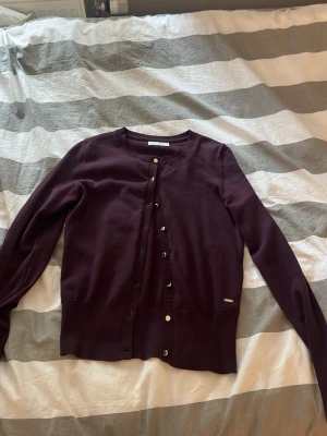 Mörklila kofta från Massimo Dutti - Stilren mörklila kofta från Massimo Dutti med rund halsringning och långa ärmar. Koftan har knäppning framtill med silvriga knappar och en liten metallbricka med logga vid nederkanten. Perfekt för lager-på-lager och enkel att matcha.