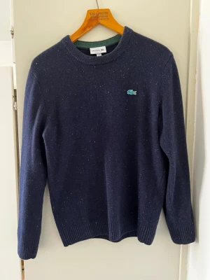 Mörkblå stickad tröja Lacoste - Mörkblå stickad tröja från Lacoste med rund halsringning och diskret melerad effekt. Tröjan har det klassiska gröna krokodilmärket broderat på bröstet och ribbade muddar vid ärmslut och nederkant. Perfekt för dig som gillar stilren och enkel design.
