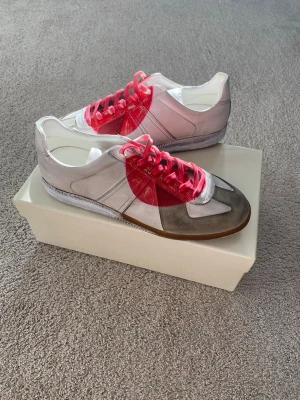Maison Margiela Japan 2006 Gats sneakers - Maison Margiela Gats sneakers i vitt och grått läder med beige mockadetaljer och genomskinliga röda paneler. Skorna har rosa snören och en klassisk låg silhuett med gummisula. Perfekt för dig som vill sticka ut med en unik och modern look.