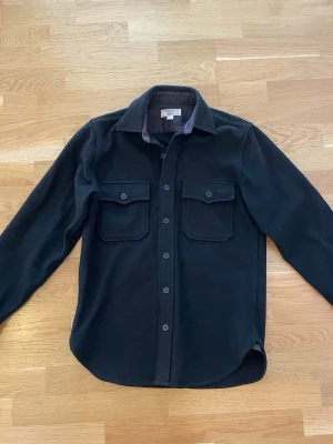 Svart overshirt från Wallace & Barnes - Stilren svart overshirt från Wallace & Barnes med klassisk krage och två stora bröstfickor med lock. Skjortan har knäppning framtill och är gjord i ett mjukt, tjockare material som passar perfekt som lager. Enkel och clean look som funkar till det mesta. Finns ett lagat hål på en av armarna men syns inte så mycket.