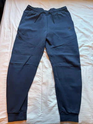 Mörkblå Nike tech mjukisbyxor - Snygga mörkblå mjukisbyxor från Nike med dragsko i midjan och ribbade muddar vid bensluten. Byxorna har en diskret Nike-logga på låret och praktiska fickor. Tillverkade i mjukt och bekvämt material, perfekta för chill eller träning.