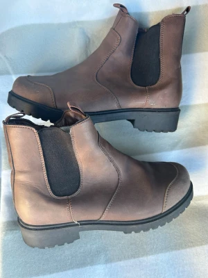 Bruna Chelsea boots med grov sula - Snygga bruna Chelsea boots i läderimitation med elastiska svarta paneler på sidorna och grov, svart sula. Klassisk siluett med dragflikar både fram och bak för enkel påtagning. Perfekta för dig som vill ha en stilren men robust look.