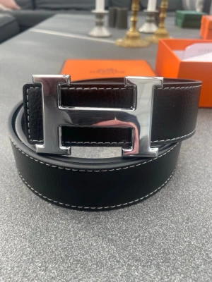 Svart H-bälte - Snyggt svart skärp med ikoniskt H-spänne i silverfärg. Skärpet har vita kontrastsömmar och är gjort i exklusivt skinn. Perfekt accessoar för att lyfta vilken outfit som helst.