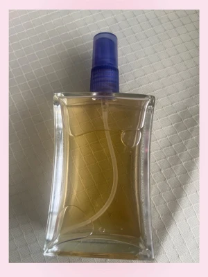 Vanilj caramel parfym - Snygg parfymflaska i genomskinligt glas med svagt böjd form och blå spraykork. Flaskan har ett elegant och modernt utseende, perfekt att ha framme på sminkbordet. Innehållet är ljusgult och flaskan har en stilren design.🩷