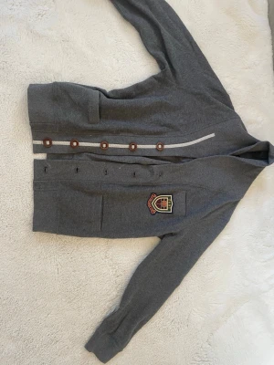 Grå kofta från Gant med emblem - Snygg grå kofta från Gant med klassiskt emblem på bröstet. Koftan har knappar framtill, två fickor och en ribbad kant. Perfekt för dig som gillar preppy stil och vill ha något som sticker ut lite extra.