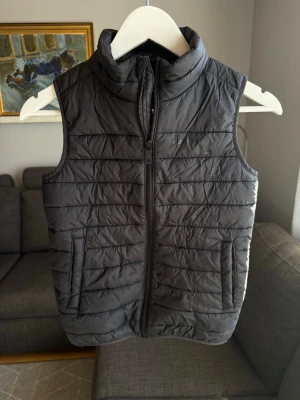 Svart quiltad dunväst - Snygg svart dunväst med hög krage och dragkedja framtill. Västen har två fickor på sidorna och ett quiltat mönster som ger en sportig look. Perfekt att slänga över en hoodie eller tröja för extra värme. Materialet är lätt och smidigt.