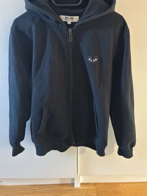 Svart hoodie från Comme des Garçons Play - Svart hoodie med dragkedja från Comme des Garçons Play. Har den ikoniska hjärtloggan med ögon på bröstet och ett stort hjärta med ögon på ryggen. Mjuk bomull, känguruficka och ribbade muddar. Perfekt för en chill och stilren look. Skriv för fler bilder!!!