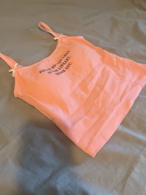 Rosa linnetopp med texttryck - Söt rosa linnetopp med tunna axelband och små rosetter vid banden. Toppen har ett svart texttryck framtill där det står 'PLEASE DO NOT CHAT IN THE LIBRARY. THANK YOU.'. Perfekt för en chill och trendig look.