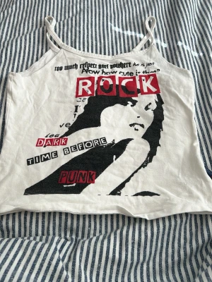 Vit croppad linne med rocktryck - Säljer ett vitt croppat linne med tunna axelband och ett stort svart och rött rock/punk-tryck framtill. Trycket har text och en grafisk bild av ett ansikte. Perfekt för dig som gillar edgy och unik stil. Y2K