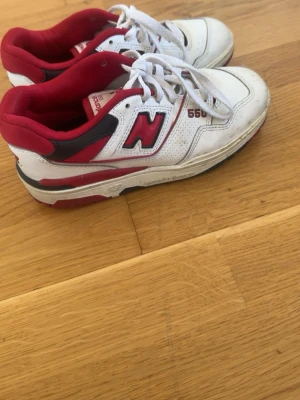 New Balance 550 vita och röda sneakers - Säljer ett par New Balance 550 sneakers i vitt läder med röda och svarta detaljer. Klassisk retrodesign med perforerade partier, snörning och den ikoniska N-loggan på sidan. Sköna och snygga skor som passar perfekt till streetwear och vardagsstil. Använde bara 2 gånger 