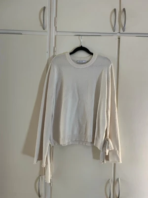 Vit stickad tröja med vida ärmar och  knytband - Säljer en stilren vit stickad tröja från NA-KD med rund halsringning och långa ärmar. Tröjan har unika knytband vid ärmsluten som ger en extra detalj. Perfekt för en enkel men trendig look.