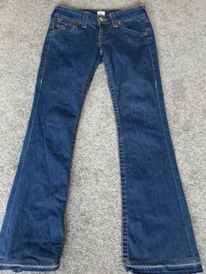 Vintage Blå bootcut jeans från True Religion - Säljer ett par vintage klassiska blå bootcut jeans från True Religion med ikonisk broderad ficka bak och snygga kontrastsömmar. Jeansen har låg midja snygg gul knapp uppsprättade där nere men annars mycket bra skick storlek w28