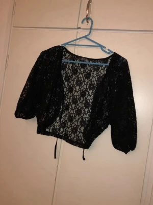 Svart spetsbolero med trekvartsärm - Säljer en svart bolero i spets med trekvartsärmar och knytband nedtill. Kort modell som är perfekt att slänga över ett linne eller klänning. Spetsmaterialet ger en snygg och trendig look.