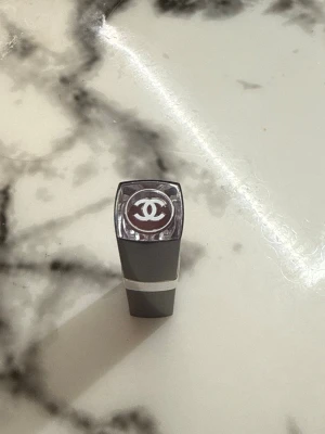 Chanel rött läppstift - Snyggt rött läppstift från Chanel med elegant svart och silverfärgat hylsa. Locket har den ikoniska Chanel-loggan i vitt. Läppstiftet har en klassisk rund form och ger en lyxig känsla till sminkväskan.
