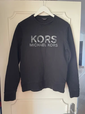Svart sweatshirt från Michael Kors i storlek S - Svart sweatshirt från Michael Kors med stor logga i grått framtill. Storlek S.Klassisk rund halsringning och ribbade muddar vid ärmslut och nederkant. Perfekt för en avslappnad och stilren look.