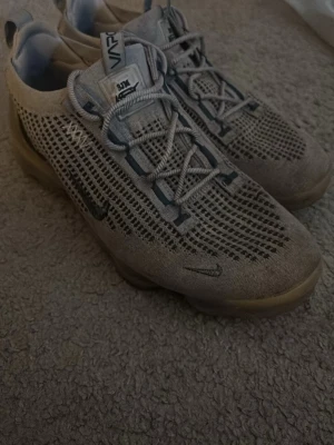 Nike Air VaporMax grå sneakers - Säljer ett par grå Nike Air VaporMax sneakers med stickad ovandel och genomskinlig sula. Skorna har små svarta Swoosh-loggor och elastiska snören. Lätta och ventilerande med modern design och coola detaljer.