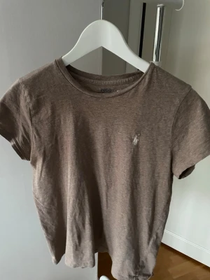 Brun t-shirt från Polo Ralph Lauren 160 cm, S - Snygg brun t-shirt från Polo Ralph Lauren med klassisk rund hals och korta ärmar. T-shirten har den ikoniska broderade loggan på bröstet och är gjord i mjuk bomull för en skön känsla. Perfekt till jeans eller shorts.