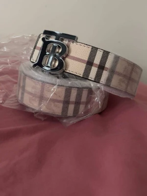 Beige rutigt bälte från Burberry - Snyggt beige bälte från Burberry med klassiskt rutigt mönster och en stor silverfärgad B-logga som spänne. Perfekt accessoar för att lyfta din outfit med en exklusiv touch. Bältet har en bred design och är lätt att matcha med olika stilar.