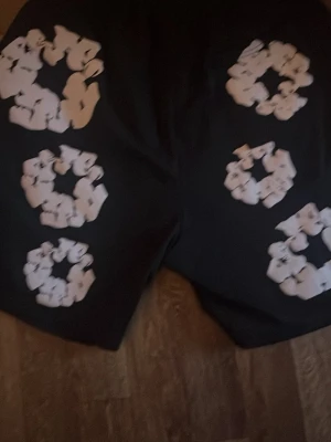 Svarta shorts från Denim Tears - Unika svarta shorts från Denim Tears med stora vita blommönster tryckta över hela plagget. Shortsen har en avslappnad passform och elastisk midja för extra komfort. Perfekta för dig som vill sticka ut med en cool och trendig stil.