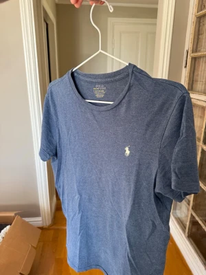 Blå t-shirt från Polo Ralph Lauren - Snygg blå t-shirt från Polo Ralph Lauren med klassisk rund hals och korta ärmar. T-shirten har den ikoniska vita polospelaren broderad på bröstet. Tillverkad i mjuk bomull för en skön känsla och enkel stil.