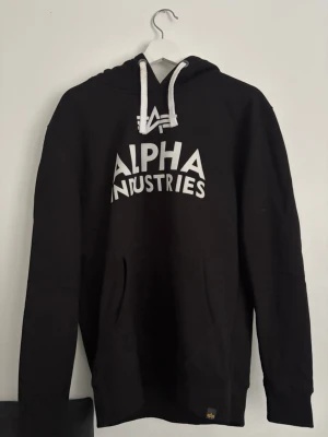 Svart Alpha Industries hoodie - Snygg svart hoodie från Alpha Industries med vit logga och text på bröstet. Klassisk känguruficka framtill, vita dragsnören i huvan och ribbade muddar vid ärmar och nederkant. Perfekt för en avslappnad och cool stil.