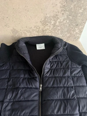 Svart dunväst från Maglia - Snygg svart dun cardigan från Maglia med quiltad framsida och stickade detaljer på krage, sidor och nederkant. Cardiganen har dragkedja framtill och en modern, sportig look. Perfekt för lager-på-lager och håller dig varm under kyliga dagar. Den är stilren och riktigt skön vill du ha fler bilder skriv mvh MW