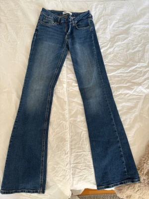 Blå bootcut jeans från Zara - Säljer ett par blå bootcut jeans från Zara med klassisk femficksdesign och snygga slitningar på låren. Jeansen har låg midja och är tillverkade i stretchigt denimtyg för skön passform. Perfekta för dig som gillar retrovibbar och vill ha ett par jeans med utsvängda ben.