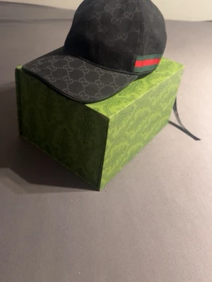 Svart Gucci keps med GG-mönster - Snygg svart keps från Gucci med det klassiska GG-monogrammet över hela kepsen. På sidan finns en grön och röd rand som ger en ikonisk touch. Kepsen har böjd skärm och justerbar rem bak. Materialet är slitstark canvas som känns lyxig och trendig.