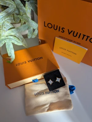 Iouis Vuitton örhänge  - Eleganta stiftörhängen i form av den ikoniska fyrbladiga blomman, presenterade med kompletta tillbehör. Design: Örhängena har en guldfärgad ram med en skimrande vit pärlemorliknande fyllning i mitten. Tillbehör: Levereras med en orange signaturask, en ljus dammpåse i tyg och en orange bärkasse. Detaljer: Inkluderar ett tillhörande infokort märkt "Monogram World Tour". Stil: En sofistikerad och tidlös accessoar som ger en lyxig touch till varje outfit.