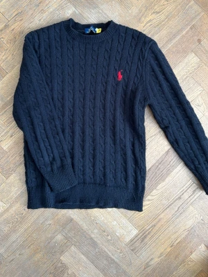 Svart kabelstickad tröja Polo Ralph Lauren - Kabelstickad svart tröja från Polo Ralph Lauren med rund halsringning och röd broderad logga på bröstet. Tröjan har långa ärmar och ribbade muddar vid ärmslut och nederkant. Perfekt för lager på lager och klassisk stil.