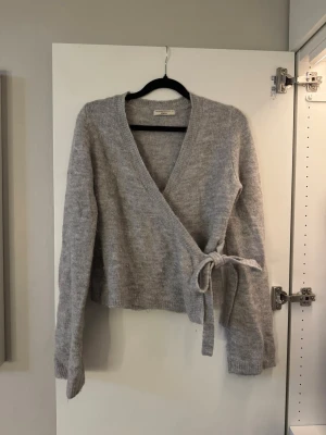 Grå omlottkofta från Gina Tricot - Mysig grå stickad omlottkofta från Gina Tricot med lite utsvängda armar och knytband i midjan. Perfekt att slänga över en topp för en avslappnad look.💛