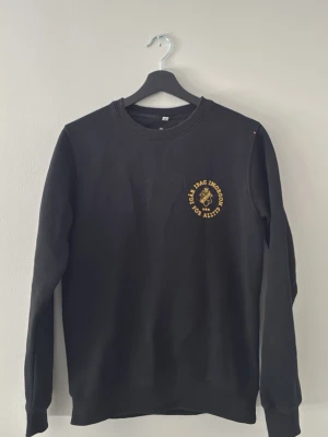 Svart sweatshirt med gul text - Svart sweatshirt med rund hals och tryck i gult på bröstet där det står 'Idag, imorgon, för alltid' runt ett lejonhuvud. Tröjan har långa ärmar och en klassisk, avslappnad passform. Perfekt för dig som gillar enkel men snygg streetwear.