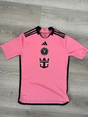 Inter Miami rosa matchtröja Adidas - Säljer en rosa matchtröja från Inter Miami CF med svarta detaljer och klubbmärke på bröstet. Tröjan har svarta Adidas-ränder på axlarna och svart logga framtill. Tillverkad i lätt och ventilerande material, perfekt för fotboll eller träning. Helt ny 100%äkta
