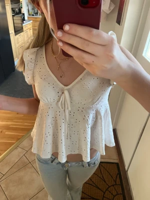 Vit broderad topp med volang - Söt vit topp med broderade detaljer och volangkant. Toppen har v-ringning, liten rosett framtill och korta ärmar med volang. Perfekt för en somrig och avslappnad stil.