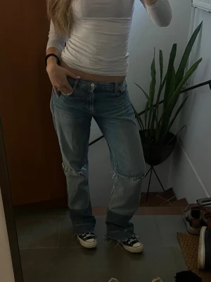 Ljusblå baggy jeans med slitna detaljer - Säljer ett par ljusblå baggy jeans med slitna detaljer vid knäna. Jeansen har klassisk femficksmodell och låg midja. Perfekta för en avslappnad och trendig look. Materialet är denim i bomull.