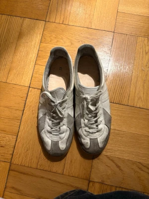 Gråvita GATs  - Säljer ett par stilrena sneakers i grått och vitt med klassisk låg profil. Skorna har ovandel i mocka och textil, vita snören och sula i gummi. Perfekta för dig som gillar retrovibbar och enkel design. Dessa är inspiration till Maison Margielas GATs. Storlek 270mm