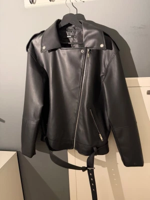 Svart skinnjacka med bälte - Snygg svart skinnjacka med asymmetrisk dragkedja, bred krage och axelklaffar. Jackan har ett bälte i midjan och dragkedjor vid ärmsluten samt en snedställd ficka framtill. Perfekt för en cool och trendig look.