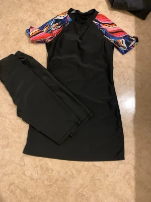 Burkini - Säljer en svart modest burkini med korta ärmar i färgglatt mönster i rosa, blått, orange och vitt. Till klänningen hör matchande svarta shorts leggings. Perfekt för stranden eller poolen. Materialet känns stretchigt och snabbtorkande.