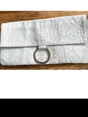 Vit kuvertväska  - Snygg vit kuvertväska i konstläder med crocomöndtrad struktur och en stor silverfärgad ringdetalj på framsidan. Väskan har en stilren och modern design som passar perfekt till olika outfits. Praktisk storlek för det viktigaste och enkel att bära i handen. 23*15 cm. Fordrad med en innerficka 