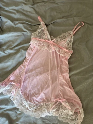 Rosa nattlinne med spetsdetaljer - Sött nattlinne i ljusrosa med tunna axelband och transparent mesh. Dekorerat med vita spetsdetaljer vid byst och nederkant samt små rosetter. Materialet är mjukt och följsamt, perfekt för en mysig kväll hemma.