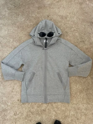 Grå hoodie med glasögon från C.P. Company - Säljer en grå hoodie från C.P. Company med ikoniska inbyggda glasögon i huvan. Hoodien har dragkedja framtill, två snedställda fickor med dragkedja och ribbade muddar. Perfekt för dig som vill sticka ut med en unik streetwear-look.