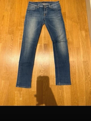 Blå raka jeans från Nudie  - Klassiska blå jeans från Levi's med raka ben och femficksmodell. Jeansen har en mellanblå tvätt med snygga slitningar och subtila kontrastsömmar. Perfekta för en avslappnad och tidlös stil.
