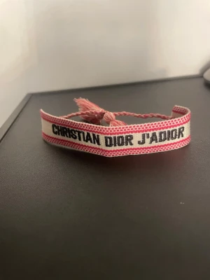Christian Dior J'Adior tygarmband - Säljer ett snyggt tygarmband från Christian Dior med texten 'J'Adior'. Armbandet är i ljusbeige med rosa detaljer och har flätade band med tofsar för knytning. Perfekt accessoar för att lyfta din outfit med en exklusiv touch.