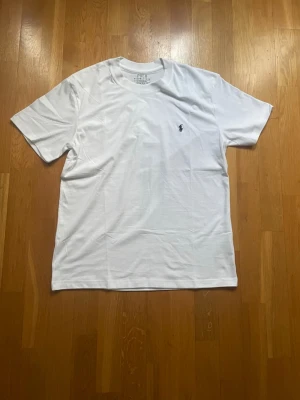 Vit t-shirt från Polo Ralph Lauren - Klassisk vit t-shirt från Polo Ralph Lauren med den ikoniska lilla loggan broderad på bröstet. T-shirten har rund hals och korta ärmar, perfekt för en clean och stilren look. Tillverkad i mjuk bomull för maximal komfort.