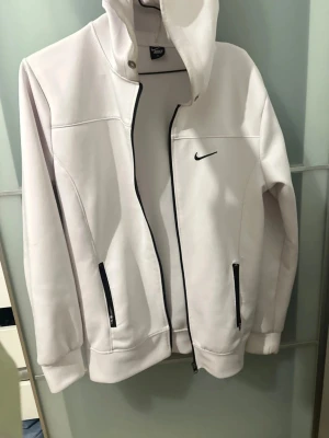 Vit hoodie från Nike med dragkedja - Snygg vit hoodie från Nike med svart dragkedja och svarta detaljer på fickorna. Klassisk Nike-logga broderad på bröstet. Hoodien har huva med tryckknappar och är tillverkad i ett mjukt, bekvämt material. Perfekt för en sportig och clean look.
